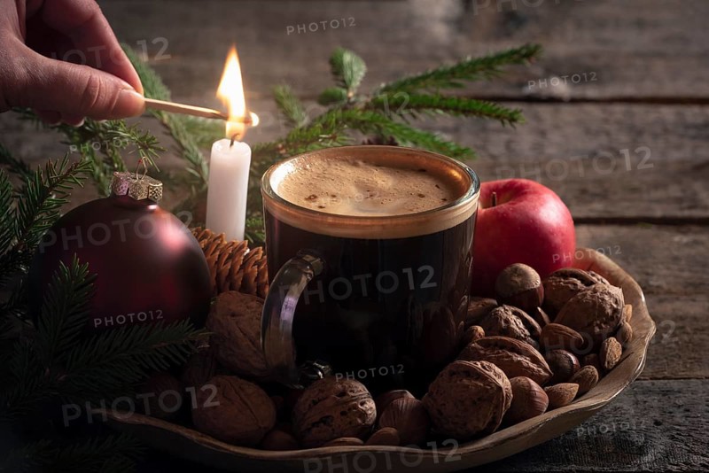 Christmas cozy atmosphere with a woman lighting a candle on a wooden platter full with nuts, apples and a cup of coffee, in low light