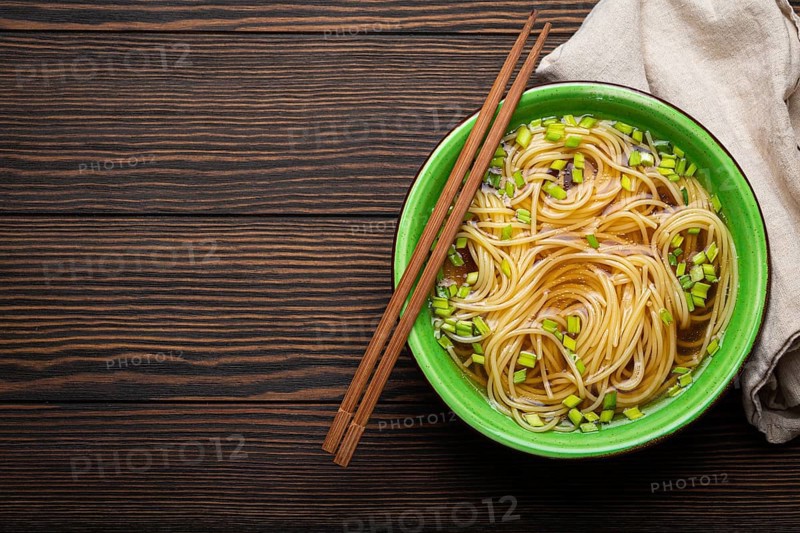 Asian noodles soup in green rustic ceramic bowl with wooden chopsticks top view on dark wooden background. Lo mein noodles with bouillon and green onion, space for text, food photography, food photography