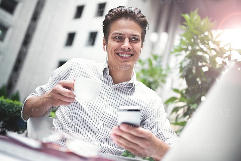 Happy handsome businessman looking at computer screen while typing something on laptop keyboard during work break outdoors, businessman with headphones looking at the notebook sitting in cafe working