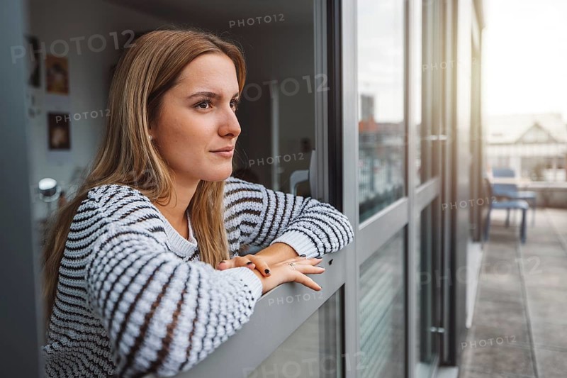 Young blonde caucasian woman looking out the window of her dorm room getting some fresh air on a cold autumn day