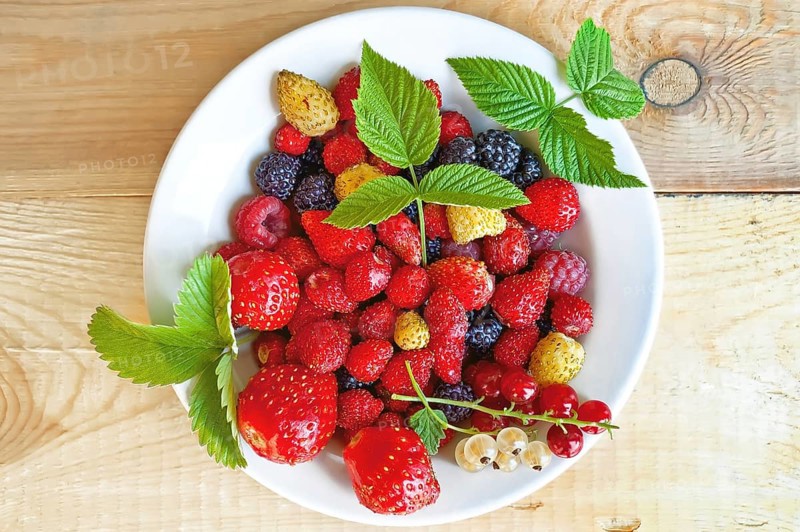 Strawberry, raspberry, blackberry, white and red currant, green leaves of strawberries and raspberry in white bowl on wooden boards