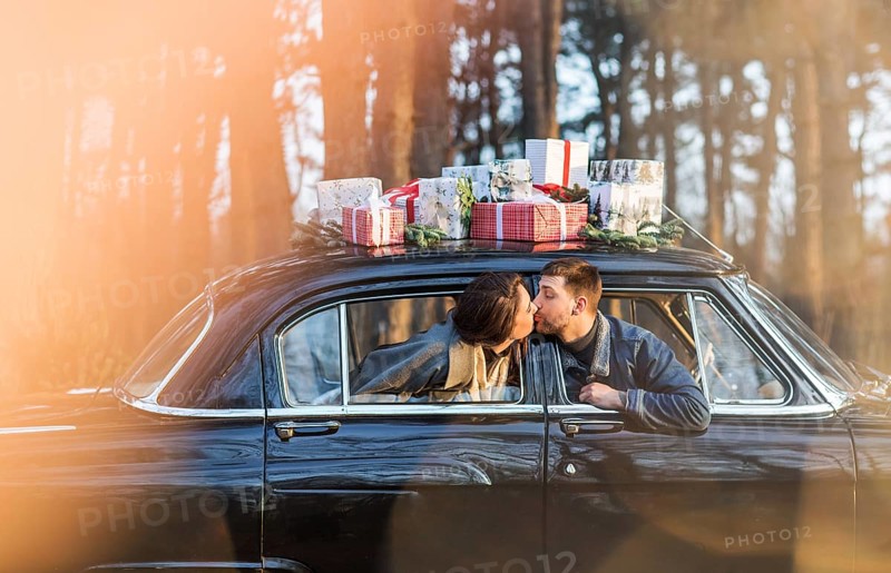 Loving man and woman sticking out of retro car windows and kissing with Christmas gifts on roof in park