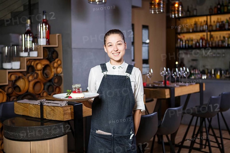 Ganson, officers, beautiful deushka with a smile and gathered hair in working form apron accepts an order in a restaurant standing near the servered wooden tables
