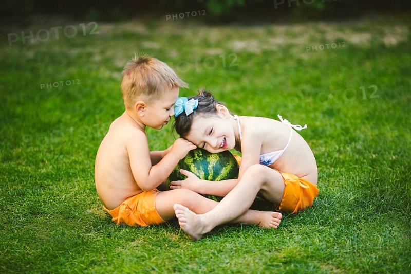 Two children, Caucasian brother and sister, sitting on green grass in backyard of house and hugging big tasty sweet watermelon berry, green striped colour. Theme healthy food and ecological product