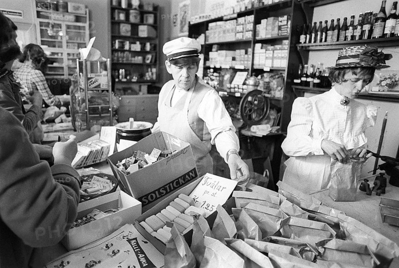 A general store museum in the 1970s. Photograph taken inside a general store, capturing its appearance from 100 years ago, are displayed for the public who shop there. The images show a nostalgic scene where customers purchase goods over the counter, served by a shopkeeper standing behind it, selling wares in a traditional, hands-on manner. The setup evokes a bygone era of commerce, blending history with the present as modern shoppers experience the same space once depicted in the century-old snapshots. Kristoffersson ref 8-22