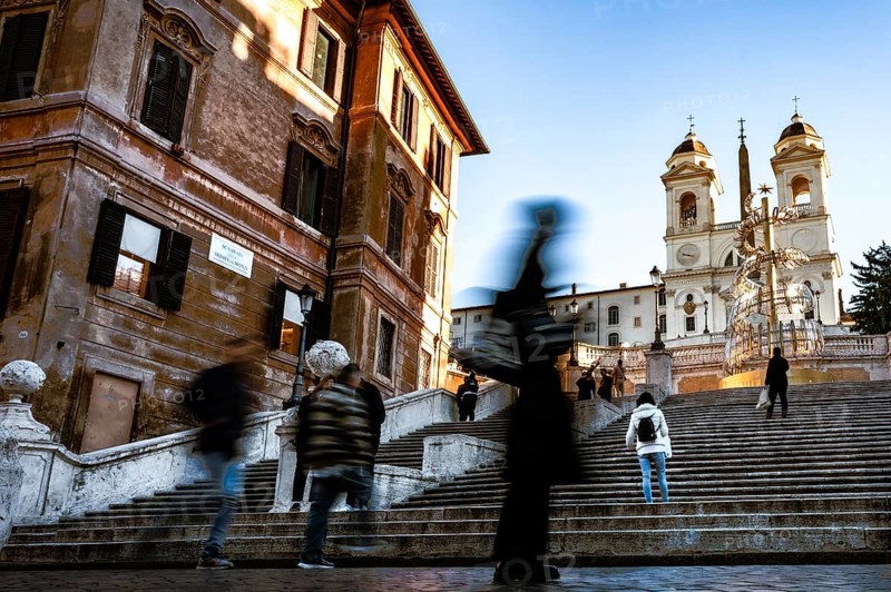 People in motion on the steps of the Spanish Steps in front of historic buildings under a blue sky, Rome