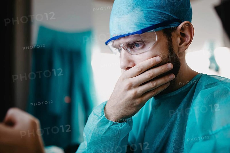 A surgeon in blue attire, appearing concerned with a hand on the mouth