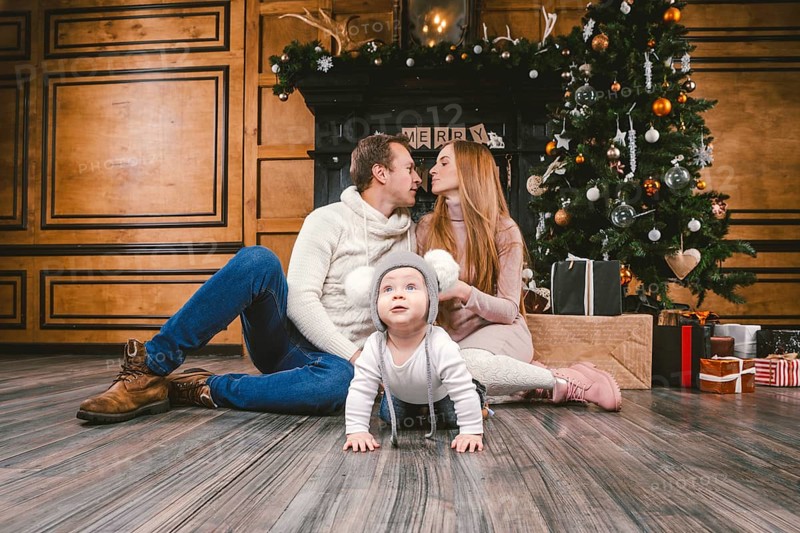 Theme family holiday New Year and Christmas. Young caucasian family mom dad son 1 year sit wooden floor near fireplace christmas tree on Christmas evening. Baby learning walk creeps background parents