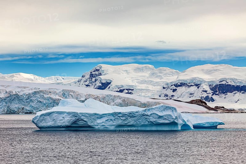 A large iceberg floats in the sea, surrounded by snowy mountains under a blue sky, iceberg in the Southern Ocean in Antarctica