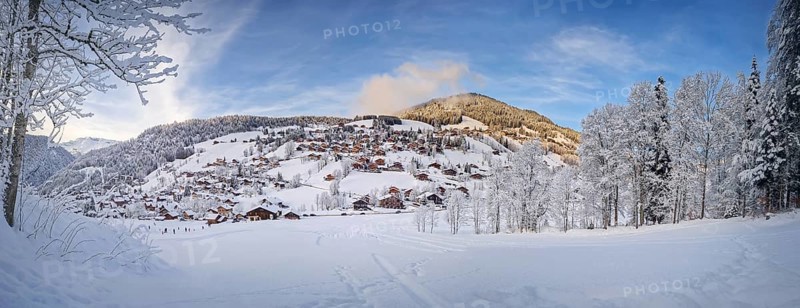 Panoramic winter scene of snow covered La Clusaz village nestled in a hilly landscape in Annecy, France. Wooden cabins are scattered across the snowy hillside, surrounded by snow laden trees