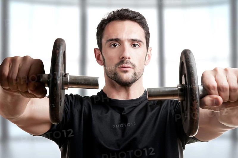 Personal Trainer doing front dumbell raises for training his deltoids in a brown background