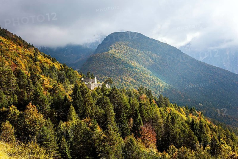Travel to Slovenia. Ruins of a medieval castle on a mountainside. The mountain road passes through yellow and orange trees and grasses. The Julian Alps