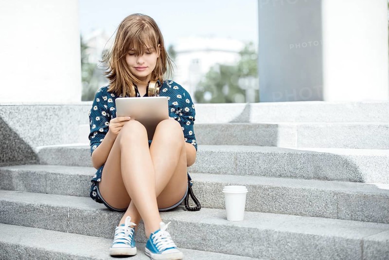 Beautiful young woman with vintage music headphones around her neck and a cup of coffee to go, surfing the internet on tablet PC and sitting on the stairs against urban city background