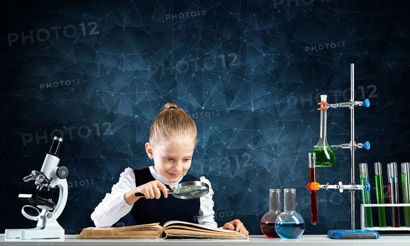 Little girl sitting at desk with magnifier and open book. Research and education in school. Happy schoolgirl in school wear carefully studying book. Clever girl reading book with magnifying glass