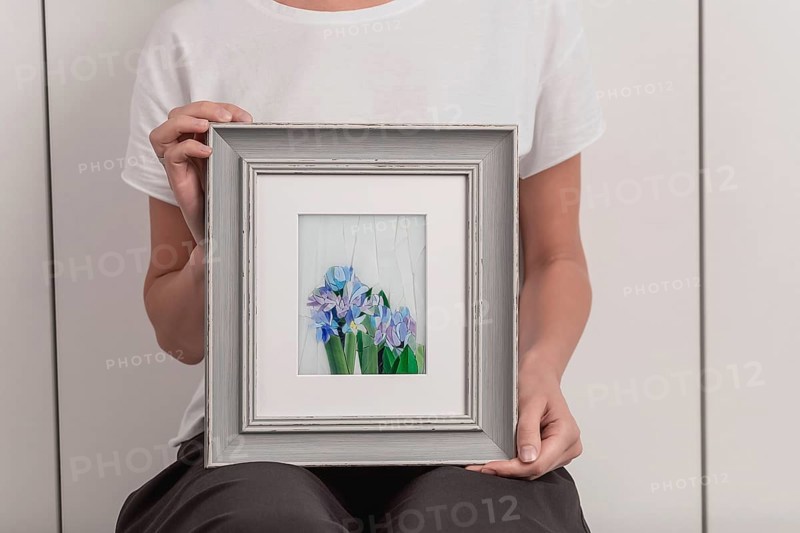 Young blonde woman in a white T-shirt is standing in a small workshop and holding in hands a mosaic picture in a frame. Close-up