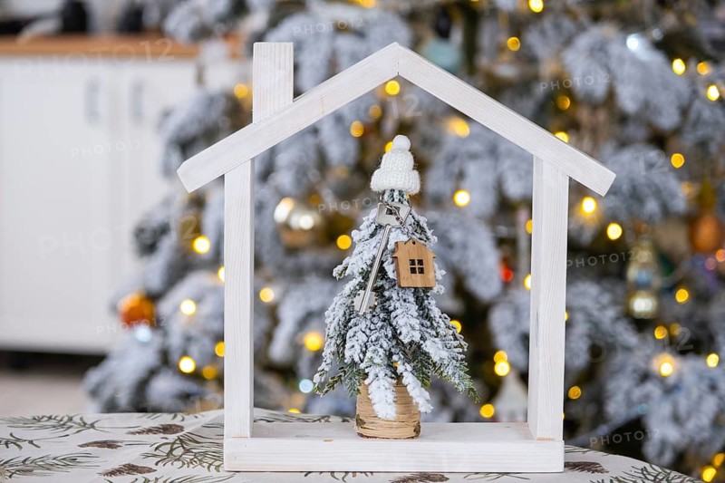 Key on Christmas tree and tiny house on cozy home with Christmas decor on table of festive white kitchen. Gift for New Year. Insurance, moving to new house, mortgage, rent and real estate