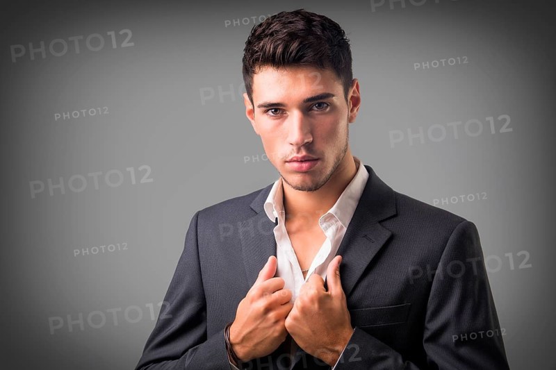 Young businessman confidently posing in front of camera, wearing business suit without neck-tie, with shirt open on neck, on dark background