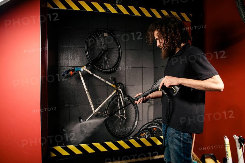 Services professional washing of a bicycle in the workshop. Close-up of hand Young Caucasian stylish man doing bicycle cleaning using automatic electric water pump. Sprays scatter from the pressure