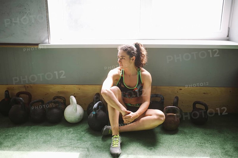 The girl is resting, taking a break during training in the gym. Sits by the window near a lot of weights