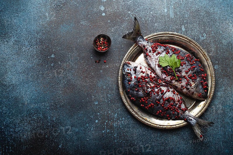 Two raw fishes dorado seasoned in pink peppercorn ready to be cooked laying on rustic metal plate top view. Healthy cooking, diet food eating concept. Dark blue concrete stone background, copy space, food photography