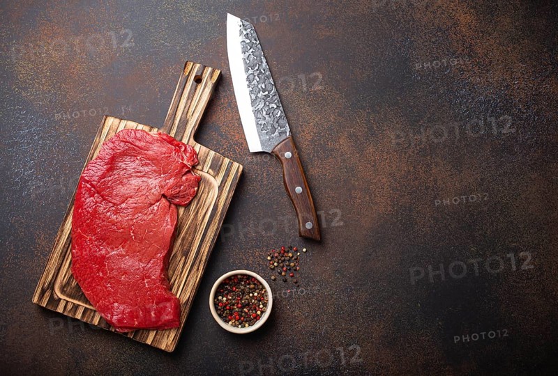 Raw uncooked top round beef steak on wooden cutting board with big kitchen knife and pepper on dark brown rustic stone background top view, cooking meat steak with copy space, food photography, food photography