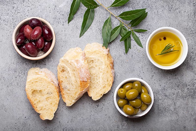 Slices of fresh ciabatta, green and brown olives, olive oil with rosemary, olive tree branches on gray concrete stone rustic background overhead, food photography, food photography