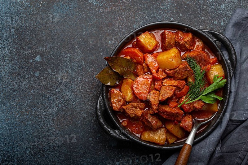 Beef meat stew with potatoes, carrot and delicious gravy in black casserole pot with bay leaves and fresh green herbs with spoon on dark rustic concrete background from above, space for text, food photography