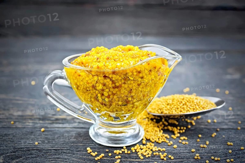 Mustard Dijon sauce in a glass sauceboat and mustard seeds in a spoon on a black wooden board background