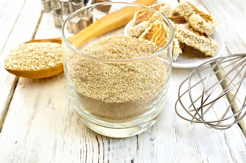 Sesame flour in a glass, a spoon with sesame seeds, cookie cutters and a mixer, biscuit on the background light wooden boards