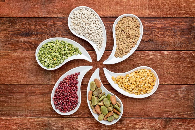 Variety of beans, lentils and pea in teardrop shaped bowls against rustic wood, top view