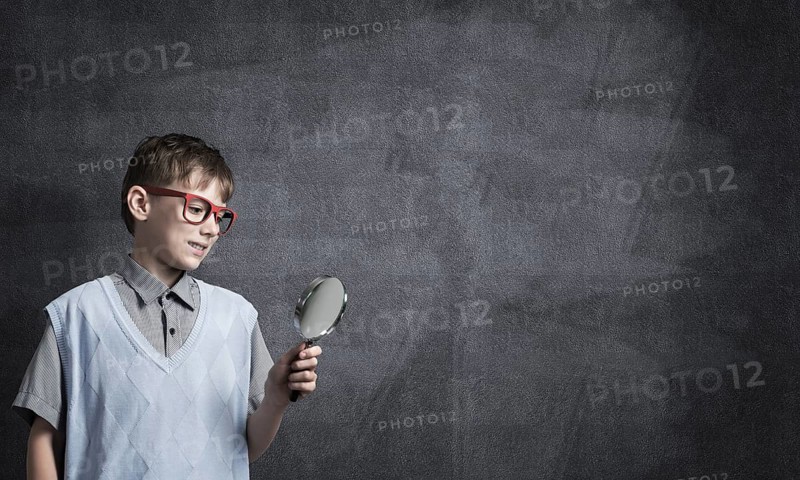 Cute boy looking through magnifying glass against blackboard background