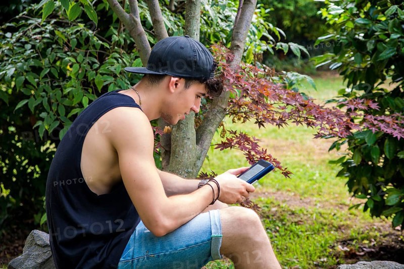 Smiling Young Handsome Man, Reading on Ebook Reader or Holding a Tablet Computer, Relaxing on Grassy Ground at Park, Leaning against Tree