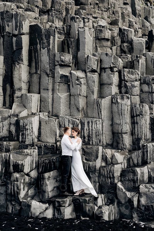 A wedding couple stands on a wall of stone pillars. The bride and groom are hugging on basalt Kekurs, on the black sandy beach of Vik. Destination Iceland wedding