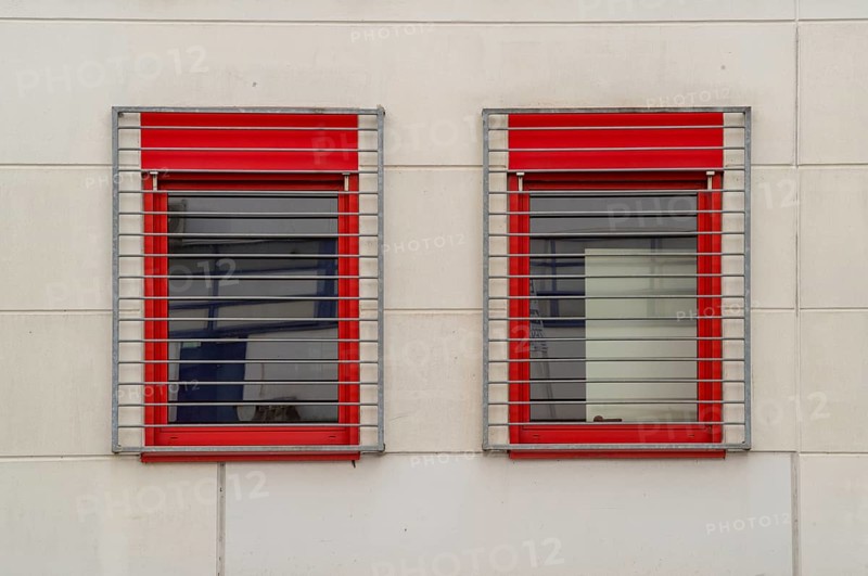 Two windows with red frames and metal grilles restrict the view outside
