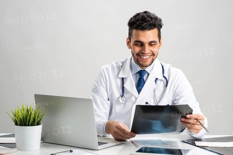 Handsome doctor looking at x-ray scan. Happy physician in white coat with stethoscope sitting at desk with laptop. Professional medical diagnosis and treatment in hospital. Medical healthcare services