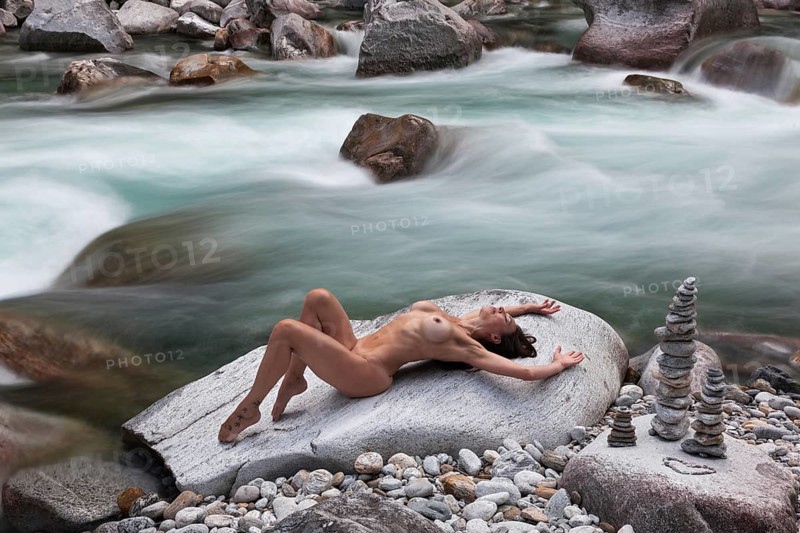 A woman in an elegantly professional pose on a rock, framed by flowing water, with pebbles stacked on top of each other to form cairns, aesthetic nude photography, outdoor shooting, Verzasca Valley, Valle Verzasca, Canton Ticino, Switzerland, Europe