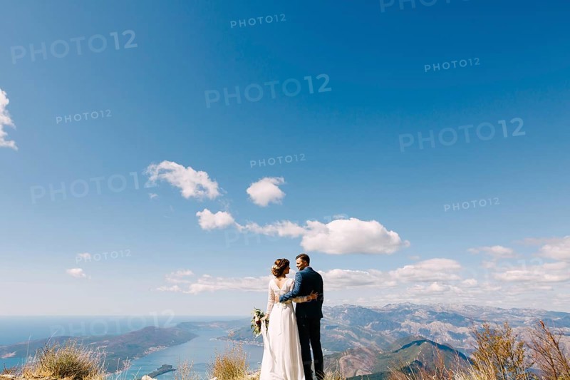 Newlyweds stand with their backs hugged against the backdrop of mountains and a bright blue sky.