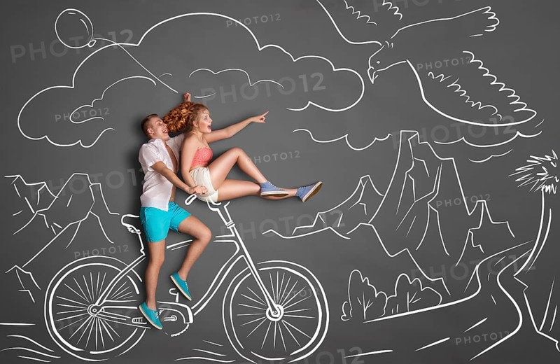 Happy Valentine's Day love story concept of a romantic couple on chalk drawings background of mountains. Male riding his girlfriend on a bicycle handlebar, female pointing at a flying eagle