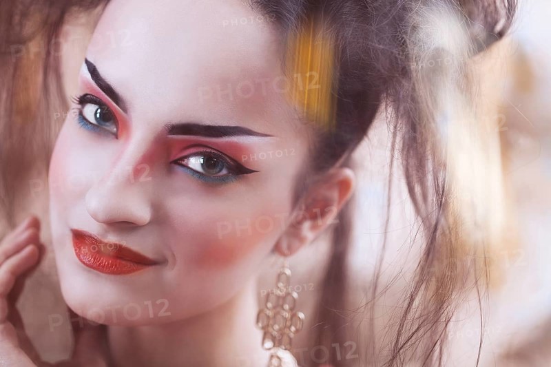 Close up stylized portrait of a Japanese geisha with bright make up standing at the window watching the rain