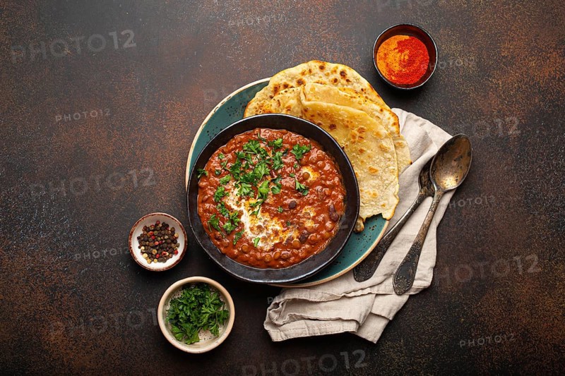 Traditional Indian Punjabi dish Dal makhani with lentils and beans in black bowl served with naan flat bread, fresh cilantro and two spoons on brown concrete rustic table top view. Space for text, food photography, food photography