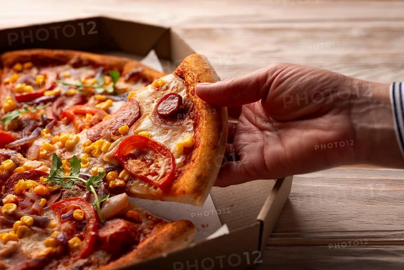 Human caucasian hand takes slice of large pizza from carton box on white wooden table