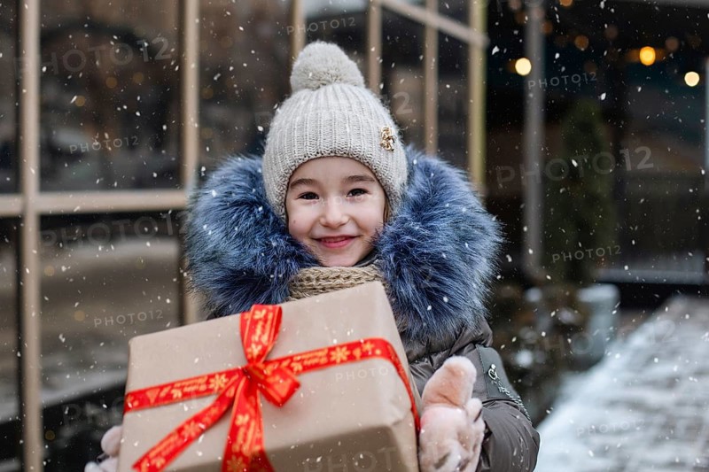 Portrait of joyful girl with a gift box for Christmas on a city street in winter with snow on a festive market with decorations and lights. Warm clothes, knitted hat, scarf and fur. Copy space