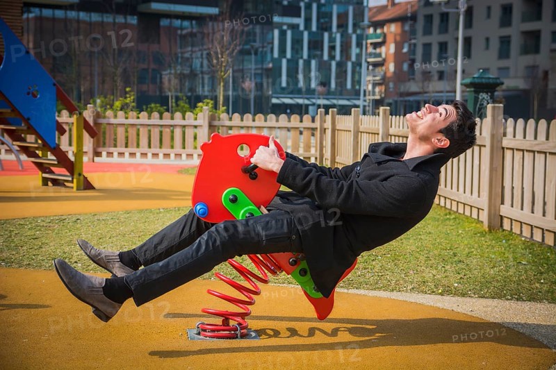 Young man reliving his childhood plying in a childrens playground riding on a colourful red spring seat with a happy smile in an urban park