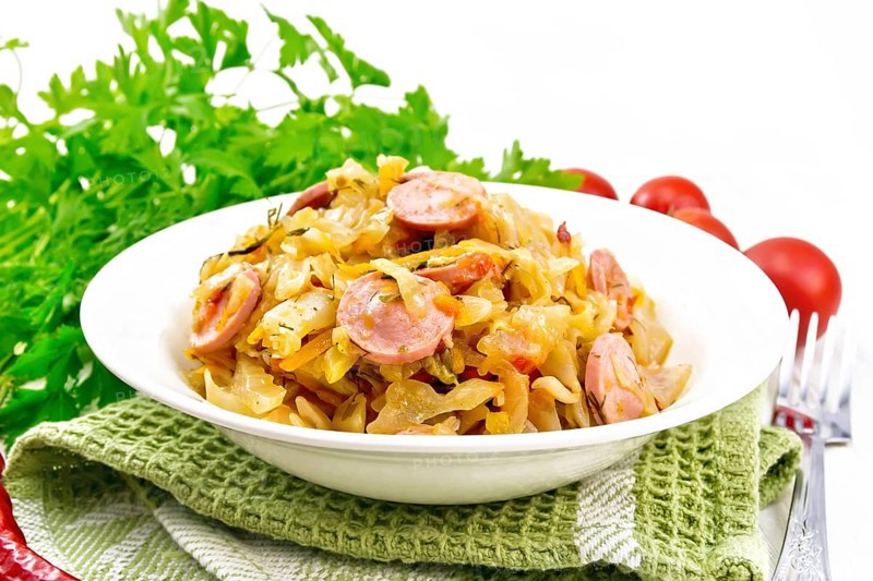 Cabbage stew with sausages in a white plate on a towel, tomatoes, parsley and a fork on the background of wooden boards