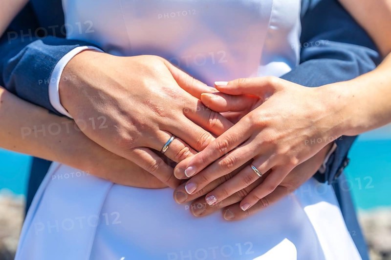 A couple is holding hands and wearing wedding rings. The woman is wearing a white dress and the man is wearing a blue suit. Scene is romantic and happy
