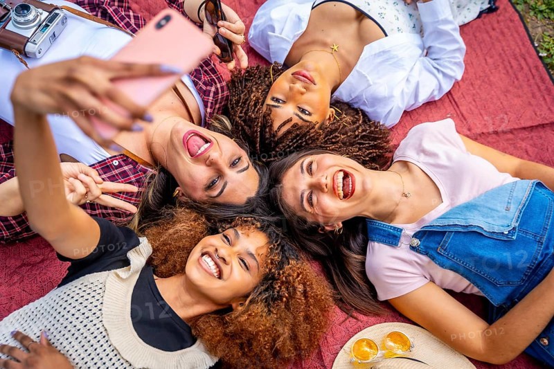 Four cheerful female friends are lying in a circle on a red blanket outdoors, taking a selfie with a smartphone