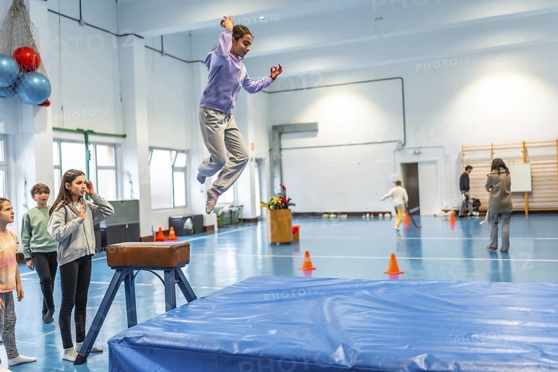 Schoolgirl practicing vaulting in a gym class, watched by her classmates and teacher, demonstrating athleticism and skill