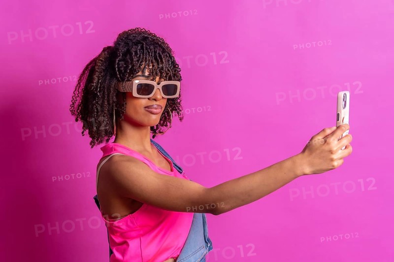 Fashionable young woman taking a selfie with smartphone, wearing sunglasses and pink top on a vibrant pink background