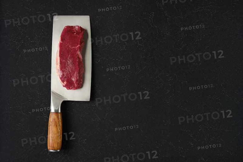 A piece of raw beef meat on a sharp cleaver with a wooden handle. The cleaver rests on a black countertop, showcasing the texture of the meat and the gleam of the blade