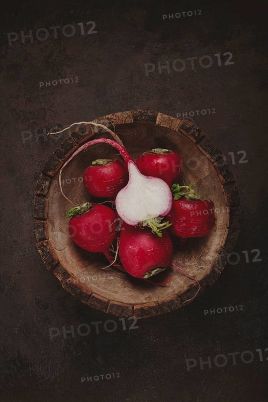 A collection of vibrant red radishes sits in a rustic wooden bowl. The contrasting dark background highlights the freshness and natural beauty of the vegetables, showcasing their green tops.